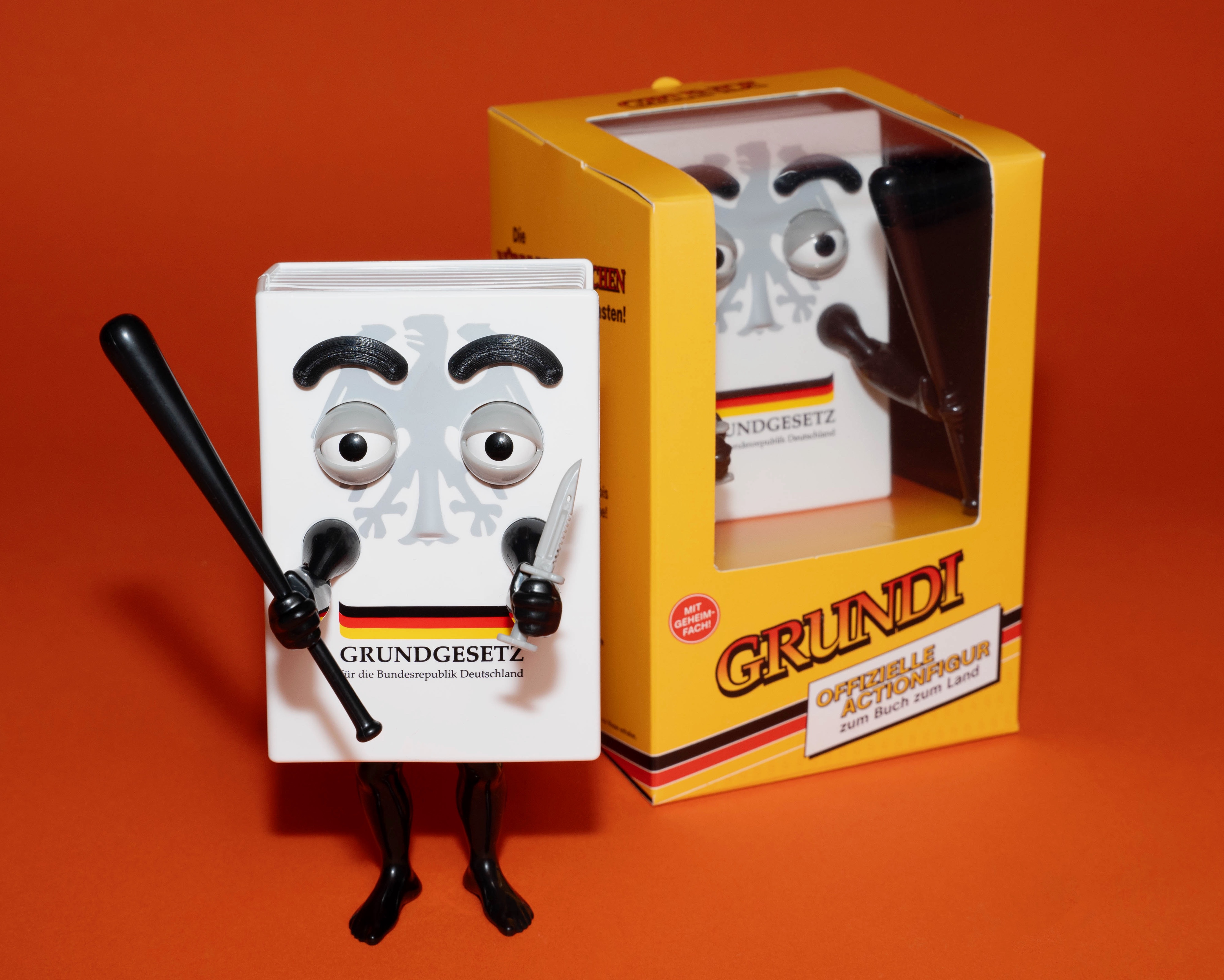 Die Grundi-Actionfigur mit Verpackung vor einem orangenen Hintergrund.